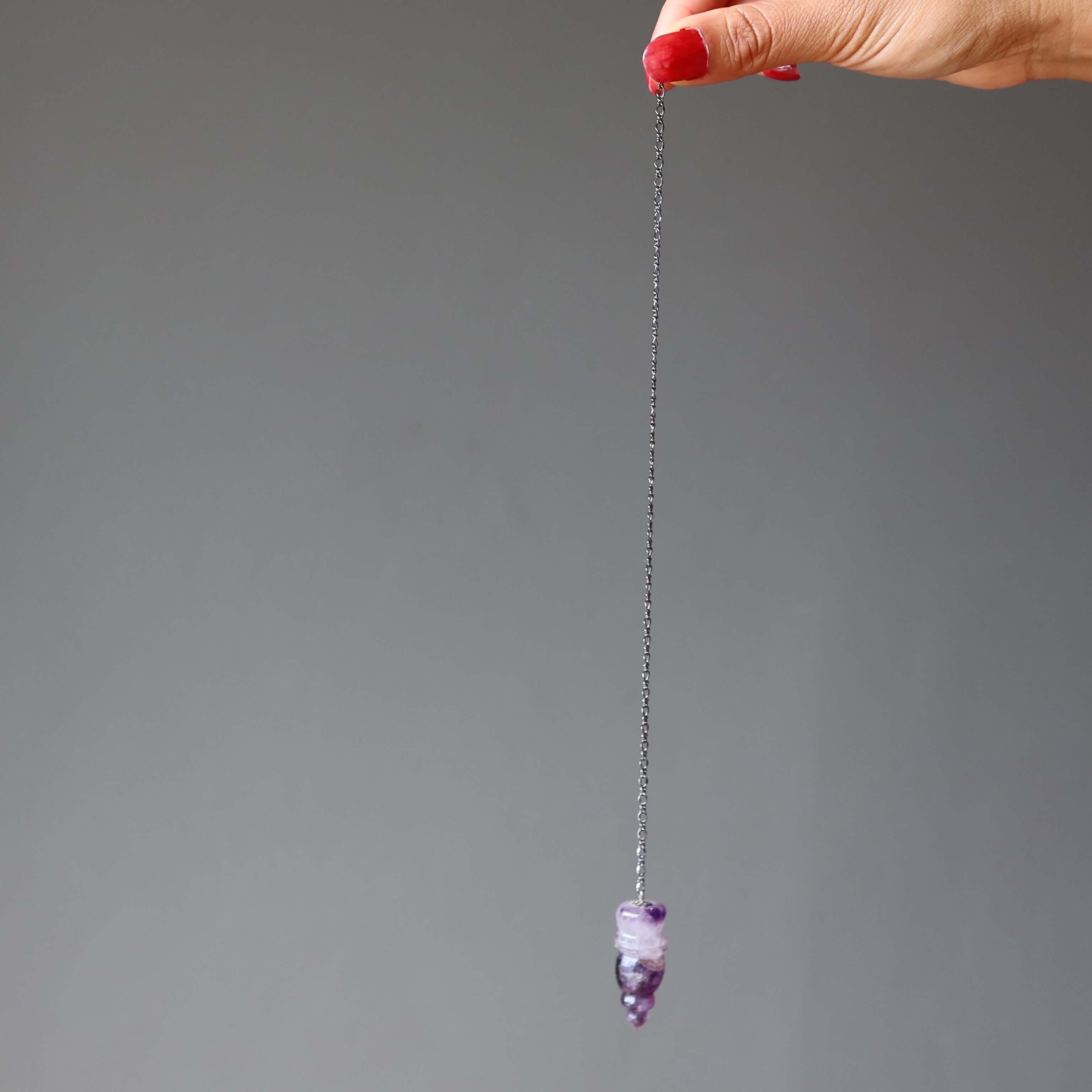 Hand holding a chain with an amethyst pendulum for dowsing, carved purple crystal point against a plain gray background