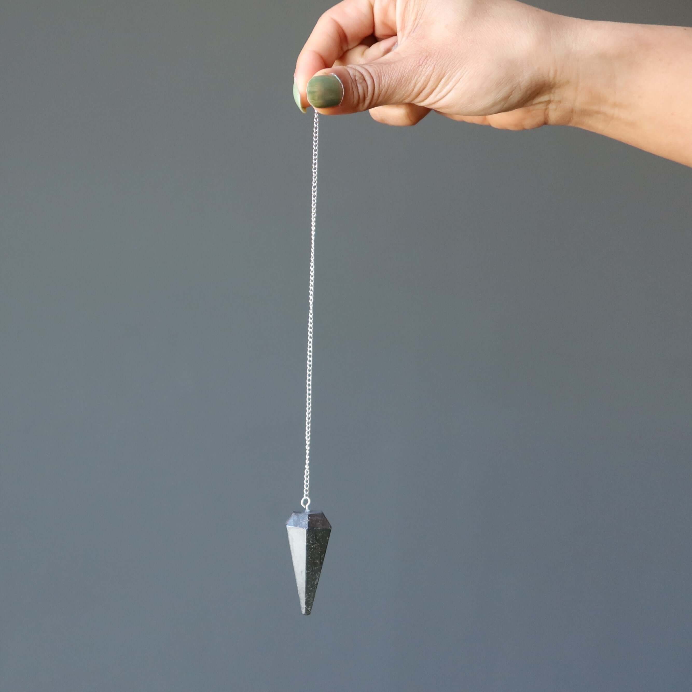 Hand holding a silver chain with a pointed Hematite pendulum for dowsing, grounding, and spiritual healing against gray background