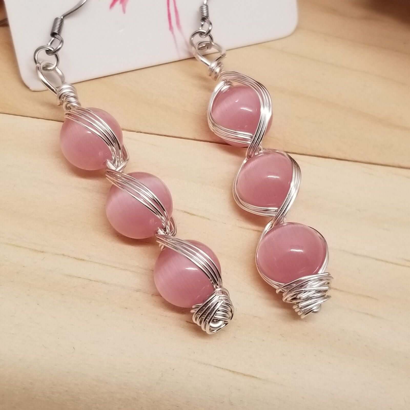 Handmade Pink Cat’s Eye Bead Earrings – Wire Wrapped Dangle Jewelry with three stacked pink beads in silver-tone wire on wood background