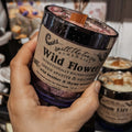 Hand holding wildflower soy wax candle in glass jar with wood wick and floral herbal design on label